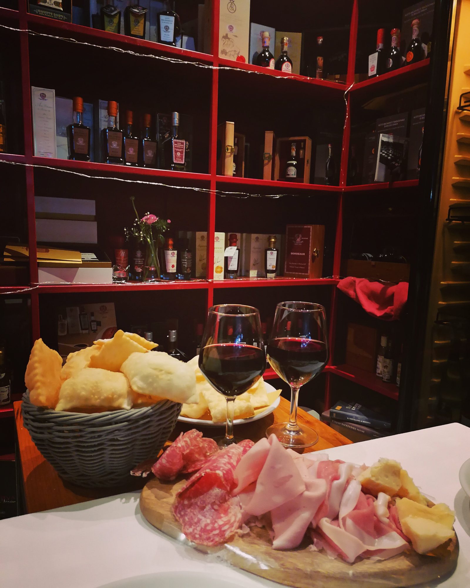 Gli scaffali rossi della bottega carichi di bottiglie, con un tagliere di salumi e gnocco fritto in primo piano